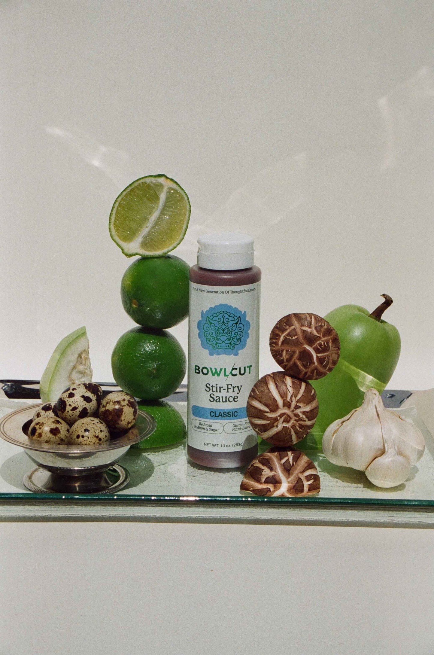 Bottle of 'Bowlcut Stir-Fry Sauce' surrounded by limes, mushrooms, and garlic on a reflective surface.
