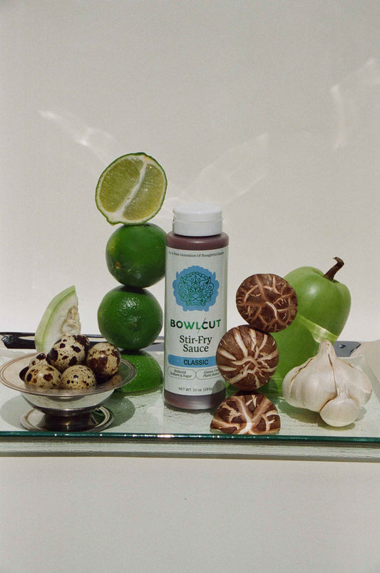 Bottle of 'Bowlcut Stir-Fry Sauce' surrounded by limes, mushrooms, and garlic on a reflective surface.